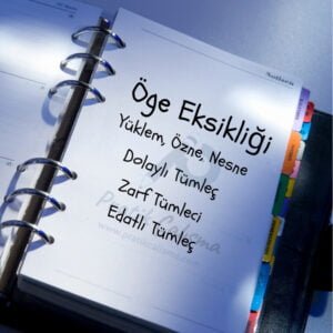 Defterin sağ üstünde "Öge Eksikliği" başlığı, aşağısında "Yüklem, Özne, Nesne, Dolaylı Tümleç, Zarf Tümleci, Edatlı Tümleç" yazıları ve bu yazıların gerisinde fon olarak "Pratik Çalışma" logosu var.
